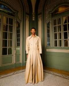 A model with medium-brown skin and short Afro hair wears a one-piece khaki outfit that features a jacket-like top attached to a flowing, floor-length skirt.