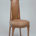 A chair made of warm, brown wood with a worn leather seat and an elongated back.