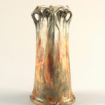 A grey and rusty colored tall and lean ceramic vase. Broad base with straight walls and a wavy flat coils on top.