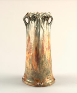 A grey and rusty colored tall and lean ceramic vase. Broad base with straight walls and a wavy flat coils on top.