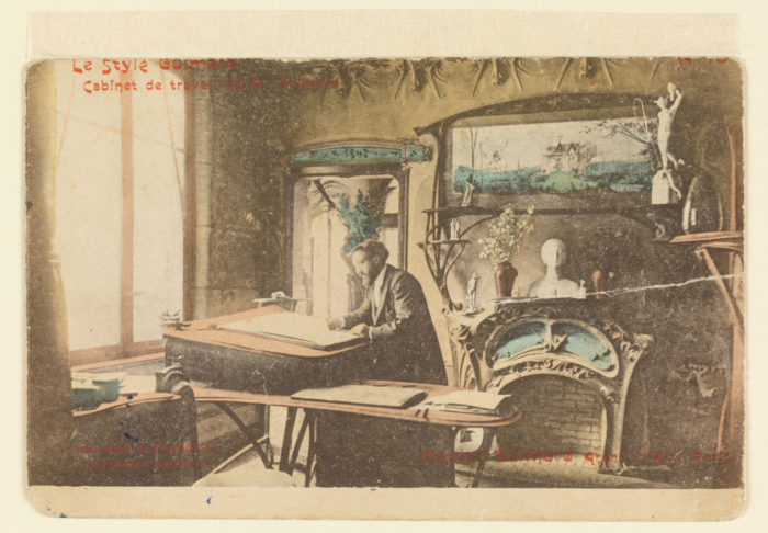 Sepia toned postcard with man sketching at drafting table with ornate fireplace in back. Top of postcard reads "Le Style Guimard"