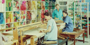 People sit working at large textile looms in front of a high wall of shelves filled to the brim with vibrant and colorful yarn and other kinds of thread; another person stands and watches their work.