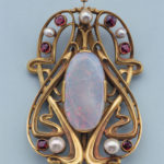 A gold buckle featuring swirling lines, a large opal stone in center, and accented with pearl and garnet .