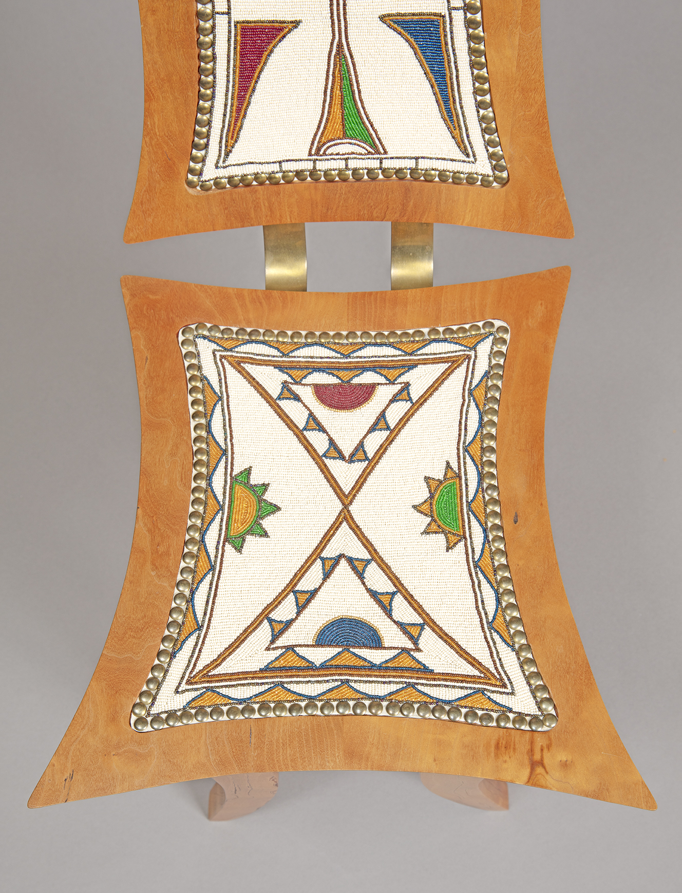 Close-up view of the back and seat of a wooden chair, rectangular shapes with concave edges and, embedded into them on deer hide, glass beads in white, brown, blue, red, green, and orange forming graphic abstract shapes.