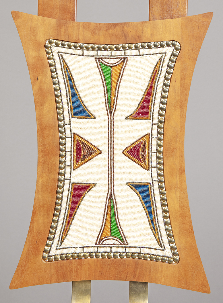 Close-up view of the back of a wooden chair, a rectangular shape with concave edges and, embedded into it on deer hide, glass beads in white, brown, blue, red, green, and orange forming graphic abstract shapes.