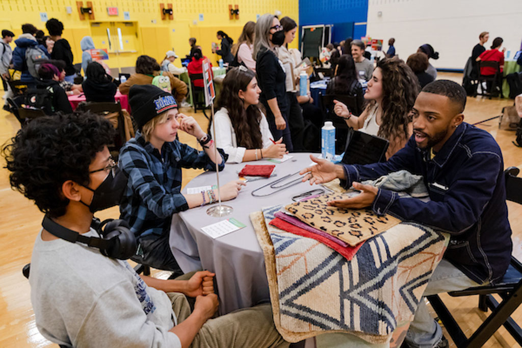 Several people sit and talk around a table in a gymnasium filled with other tables and people. Piled on this table are fabric samples with different patterns.