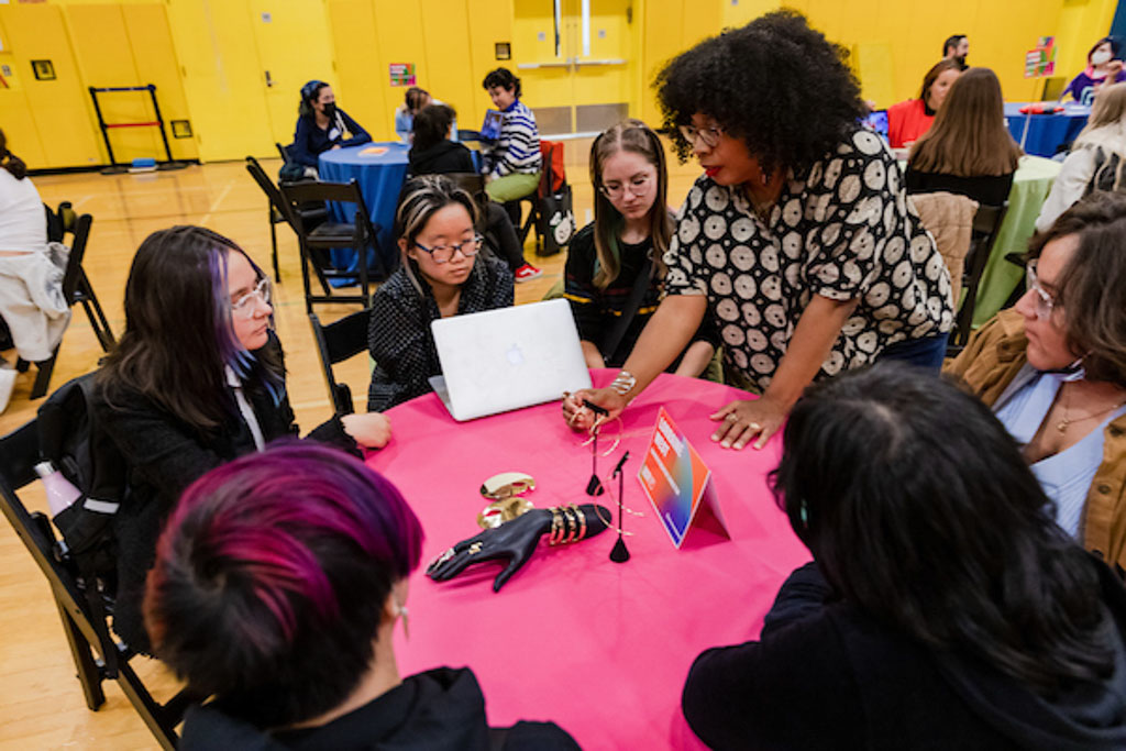 A designer stands and leans over a table to demonstrate something to a circle of seated students.