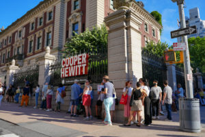 A line of people wraps around the block in front of Cooper Hewitt, a regal mansion surrounded by greenery and a stone and wrought iron fence.