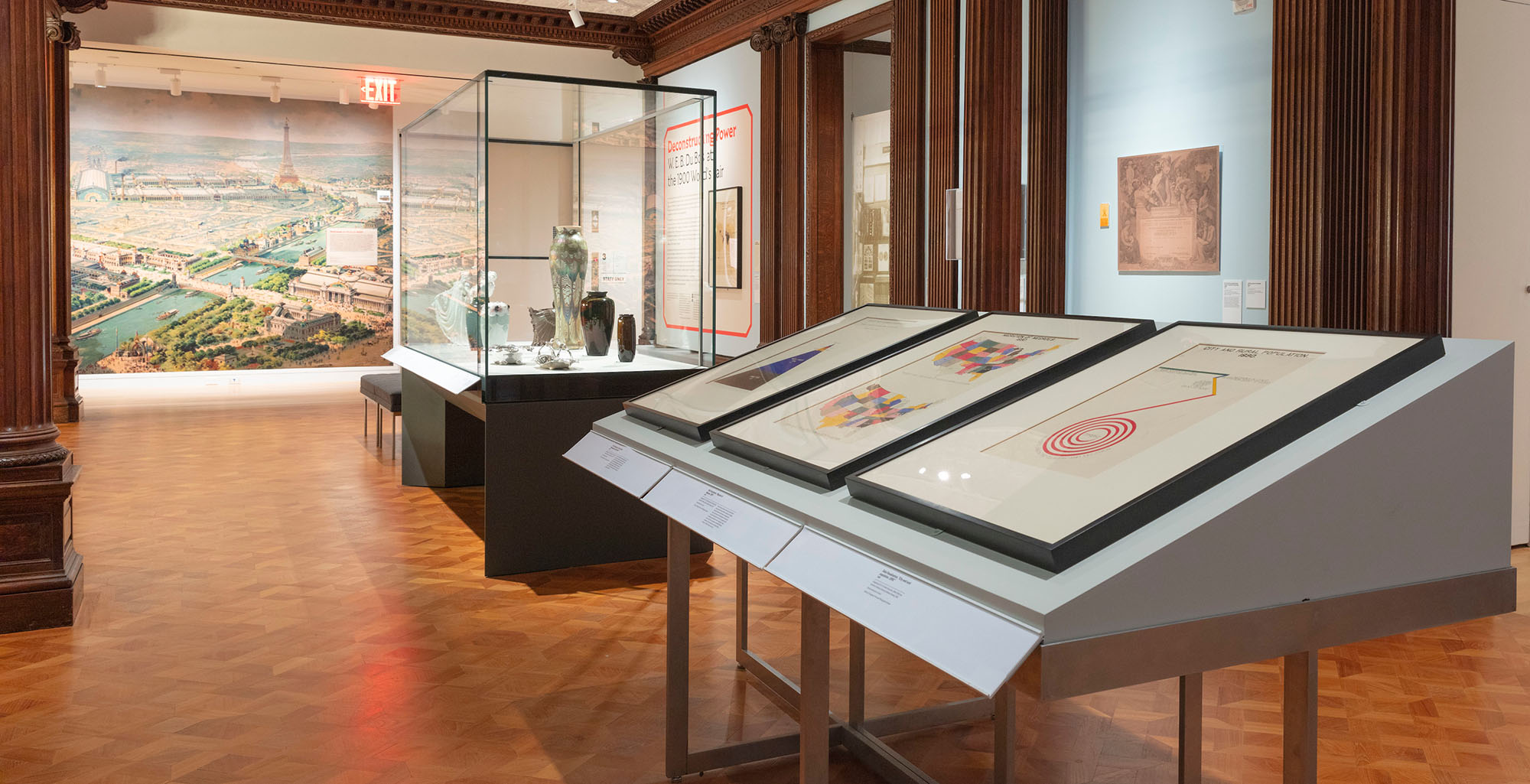 View of exhibition space with three framed artworks on a slanted table in foreground and colorful vases in a glass vitrine in background.