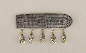 Silver, oblong-shaped brooch, with a straight side on the left, resembling the deck of a trade ship viewed from above. The deck is densely packed with silver stick figures. Five gold heads in profile hang from the bottom.