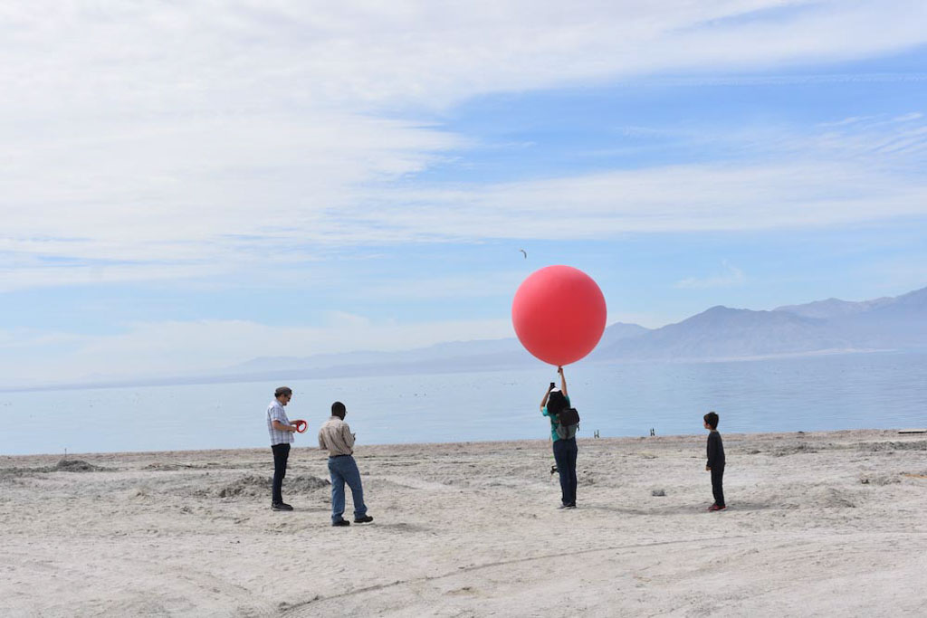 A person holds up a massive red balloon on an empty beach with a few other people gathered around.