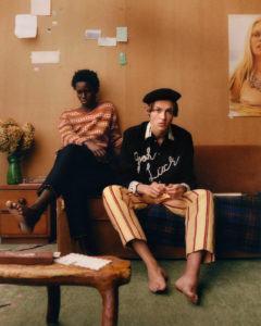 Two barefoot models pose on a couch; one wears a black sweater that says [Good Luck] in white stitched script on the front along with pale yellow pants with red pinstripes. The other wears black pants and a sweater with horizontal stripes in the same shades of yellow and red.