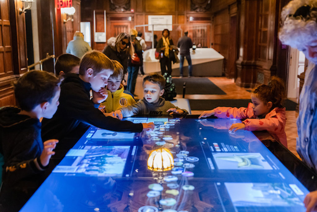 Several children gather around a touch screen interactive table in the Great Hall of Cooper Hewitt.