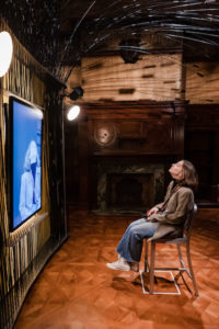 A person sits with their head tilted back on a chair in an otherwise empty wood-paneled room. They face a large screen playing a live feed of themself.