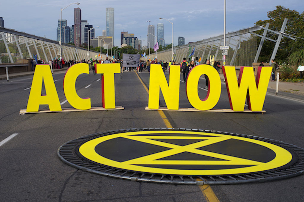 Bold, yellow letters stand in the middle of a highway spelling out [Act Now] behind a large yellow circle containing an X.