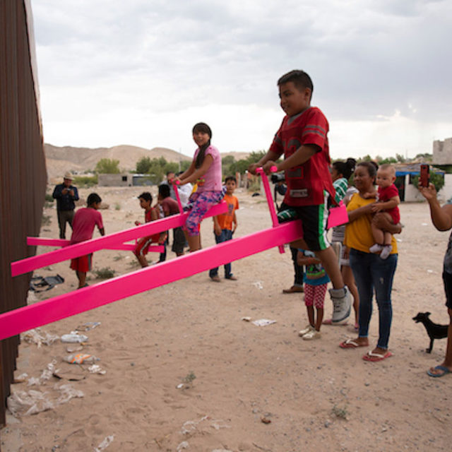 A few brown-skinned children sit and smile on hot pink teeter-totters, the other end of the teeter-totters not visible beyond the tall metal fence that they run through.