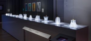 A series of glass vessels displayed in a gallery and lit from above to create a variety of dramatic light patterns on the platform.
