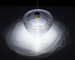 A bowl-shaped glass vessel lit from above causing dramatic and intricate patterns of light and shadow to be cast around it, uneven concentric circles of lessen intensity as the light travels farther from the light source and vessel.