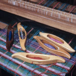 Five oblong wooden loom shuttles scattered across a fabric in progress in a loom.