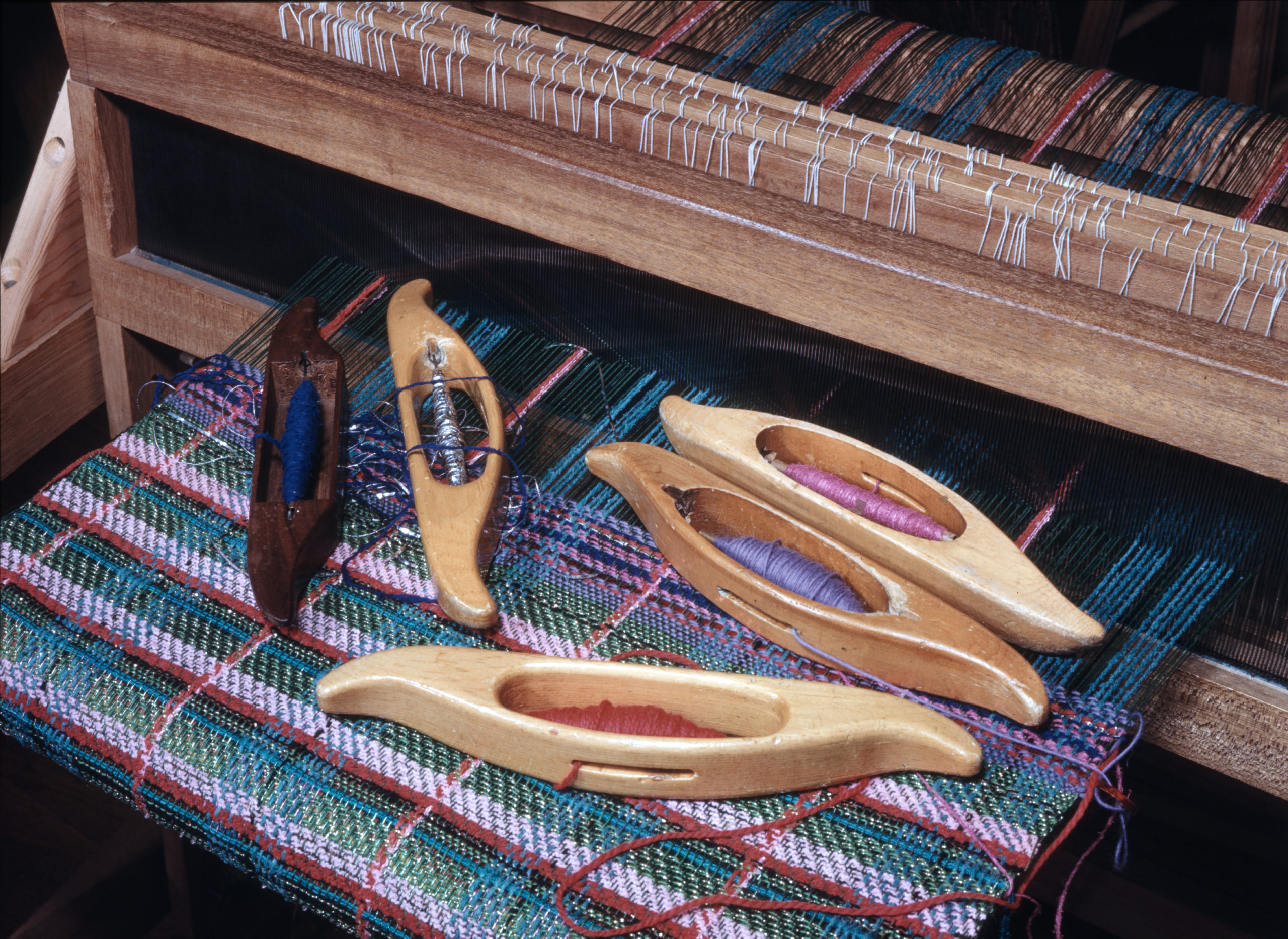 Five oblong wooden loom shuttles scattered across a fabric in progress in a loom.