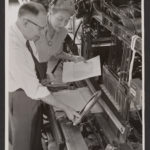 A black and white photograph of a white man and woman standing over a loom reviewing a weaving pattern.