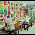 Three weavers sit at large wooden looms in front of a wall filled with floor-to-ceiling shelves of colorful yarn and other threads.