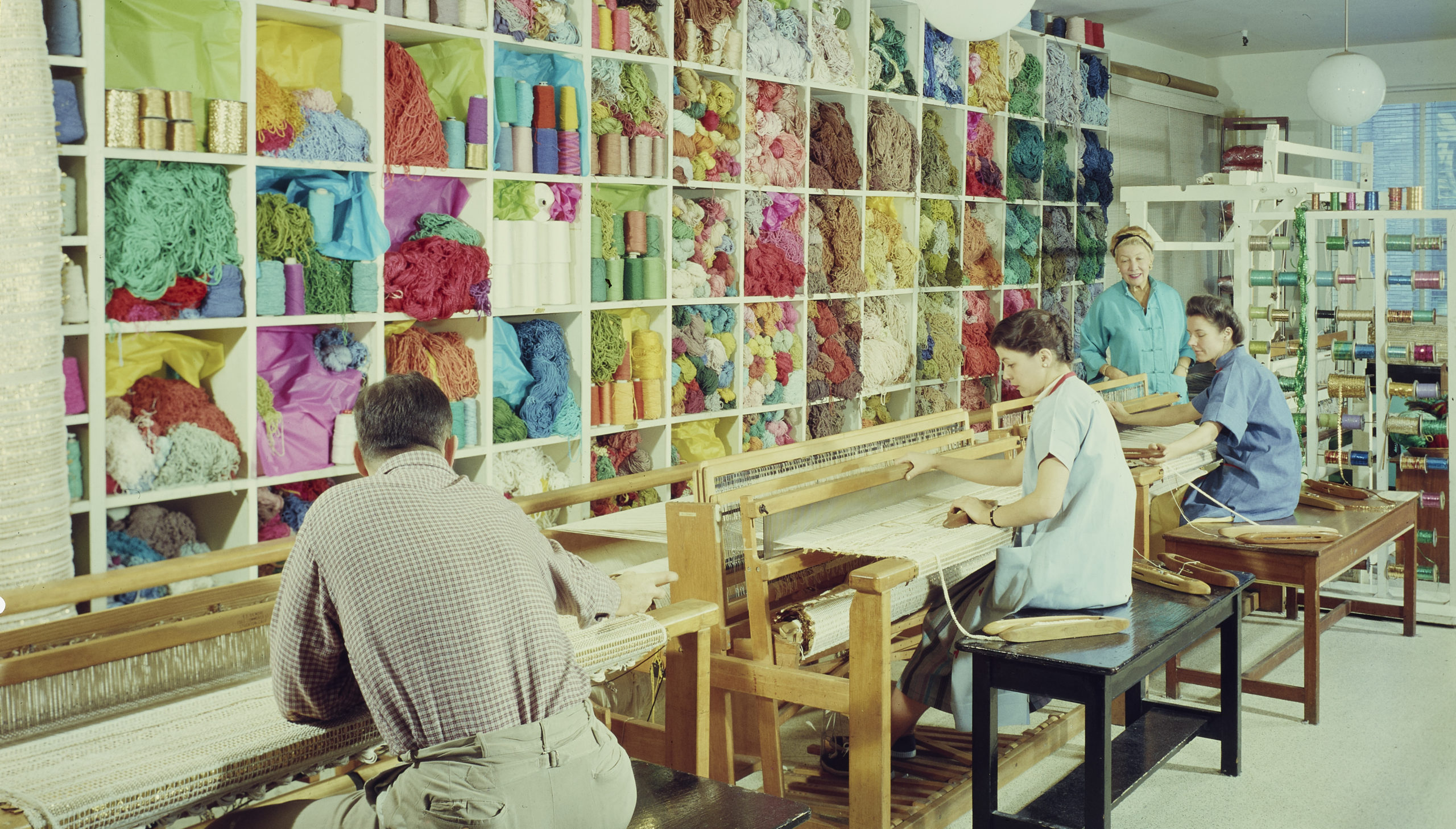 People sit working at large textile looms in front of a high wall of shelves filled to the brim with vibrant and colorful yarn and other kinds of thread; another person stands and watches their work.