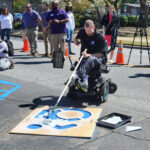 A white man in a power wheelchair uses a paint roller to apply white paint to a large accessible icon stencil on the ground.