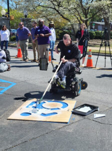 A white man in a power wheelchair uses a paint roller to apply white paint to a large accessible icon stencil on the ground.