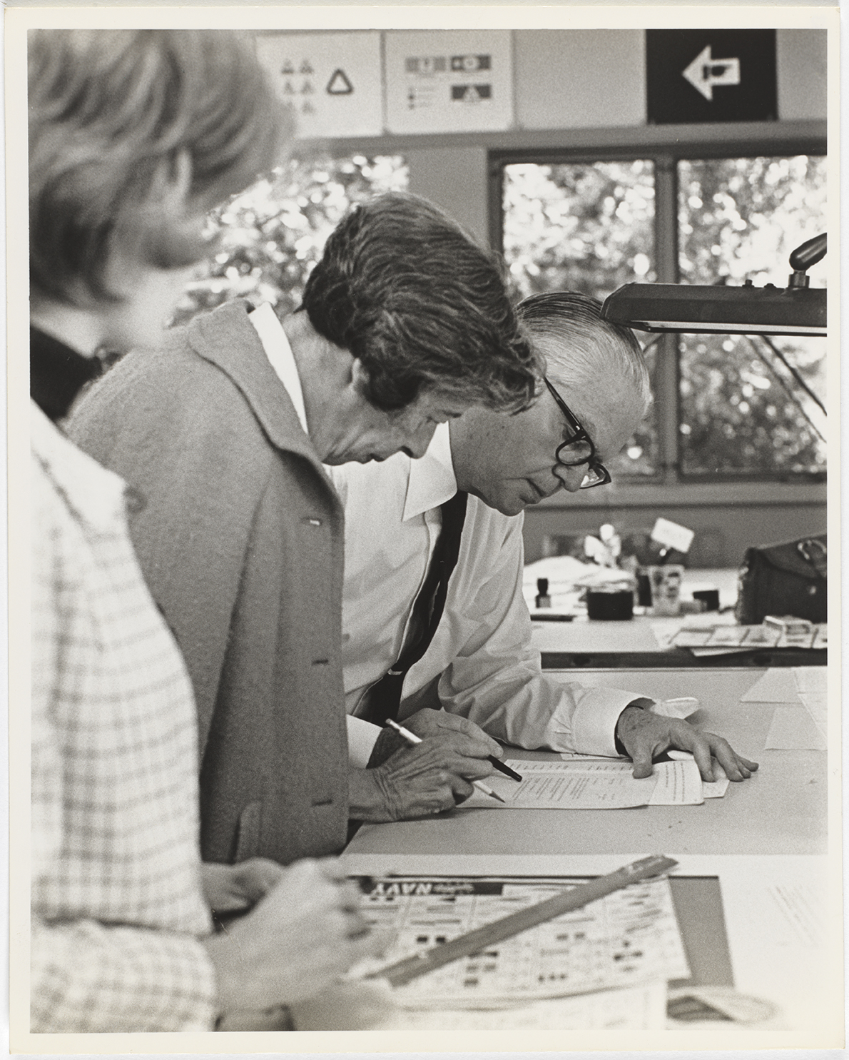 Black-and-white photograph of three people standing around a table. A man wearing horn-rimmed reads and points to a sheet of paper.