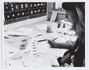 A woman with shoulder-length hair sits at a desk that is cover in papers and materials related to graphics symbols of the human form.