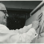 A black and white photo of Henry Dreyfuss, a white man with salt and pepper hair, sketching a large symbol on a poster in front of him.