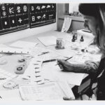 A black and white photo of a white woman looking down at a desk scattered with papers depicting various symbols.