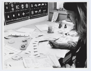 A black and white photo of a white woman looking down at a desk scattered with papers depicting various symbols.