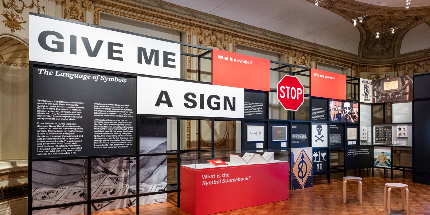 Installation view of exhibition with large sign reading "Give Me a Sign," alongside objects, including a STOP sign and poison symbol.