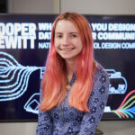 Photo portrait of a young woman smiling, standing in front of the Cooper Hewitt National High School Design Competition sign, with light pink long hair wearing a blue long-sleeved dress with flowers.