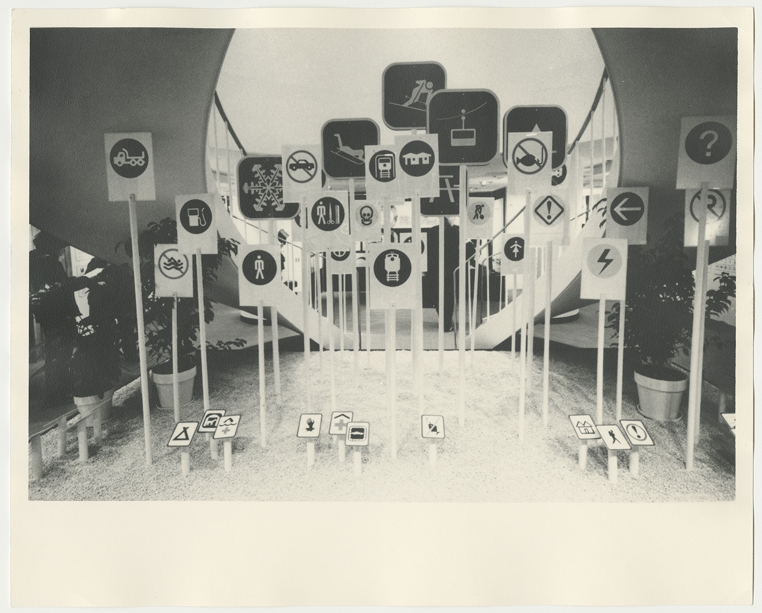 Black-and-white photograph of an exhibition featuring sticks holding up large cards depicting various symbols for communication.