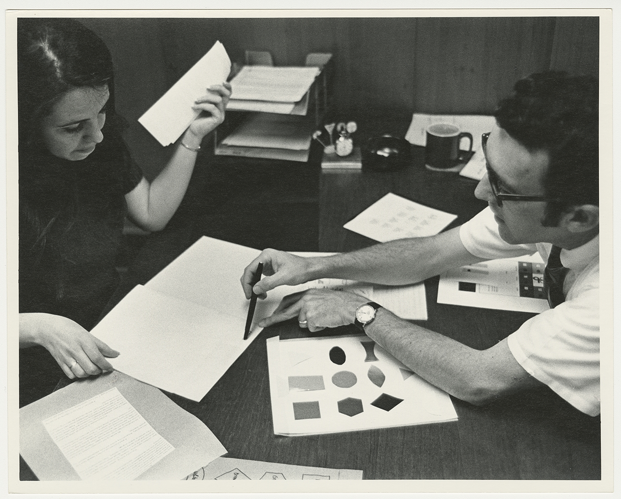 Black-and-white photograph of two people working together at a desk. Between them are papers, some containing shapes and symbols in a variety of color tones.