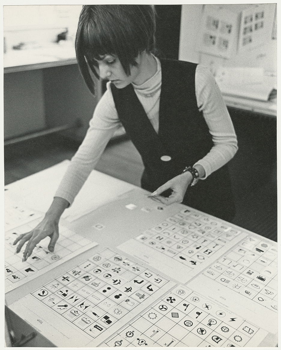 Black-and-white photograph of a woman with short hair wearing a vest leaning over a table covered papers with grids containing black-and-white graphic symbols.