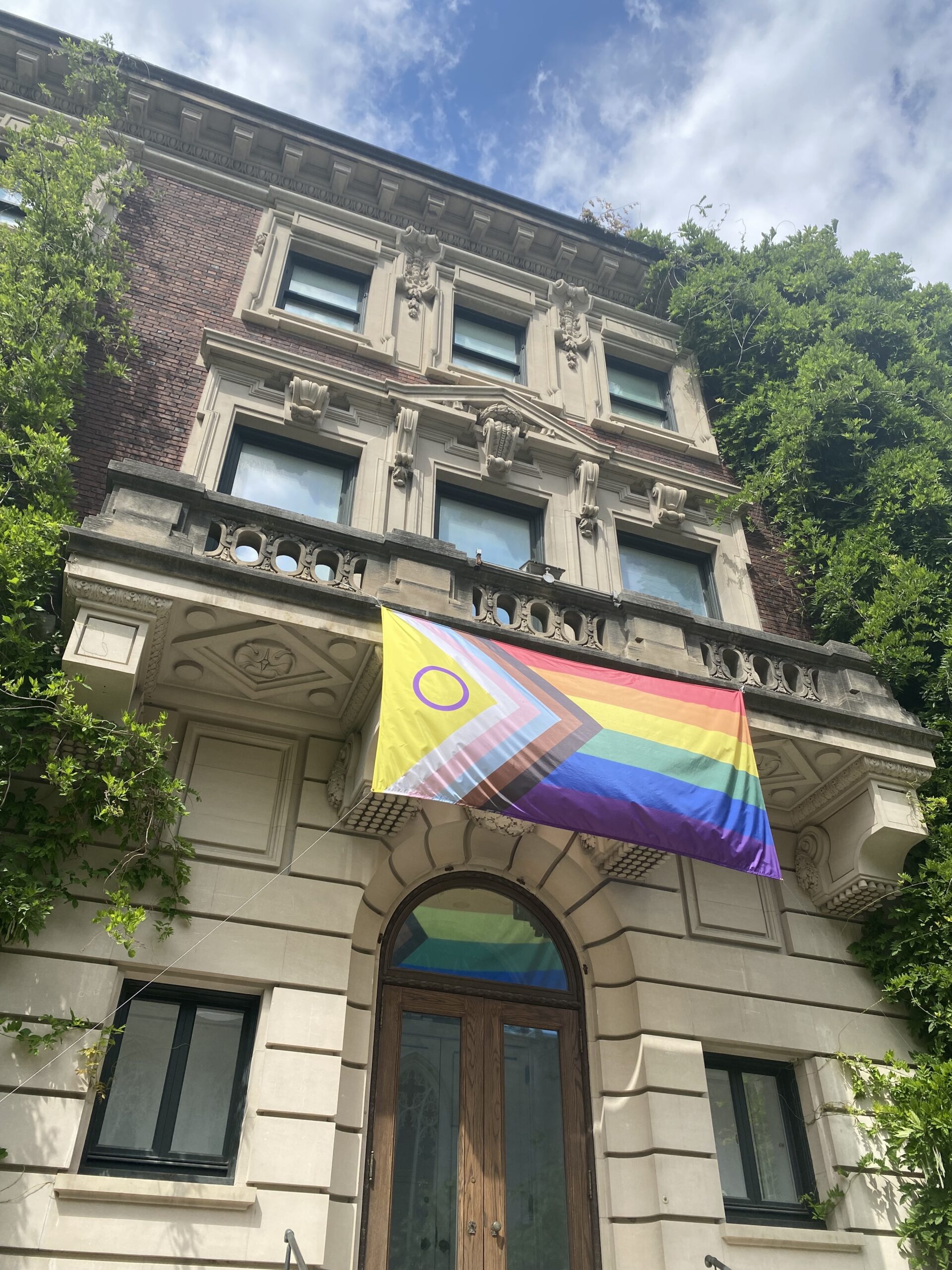 Close-up view of the Pride flag, of many stripes and colors, hanging from the balcony of an ornate mansion with foliage covering it.