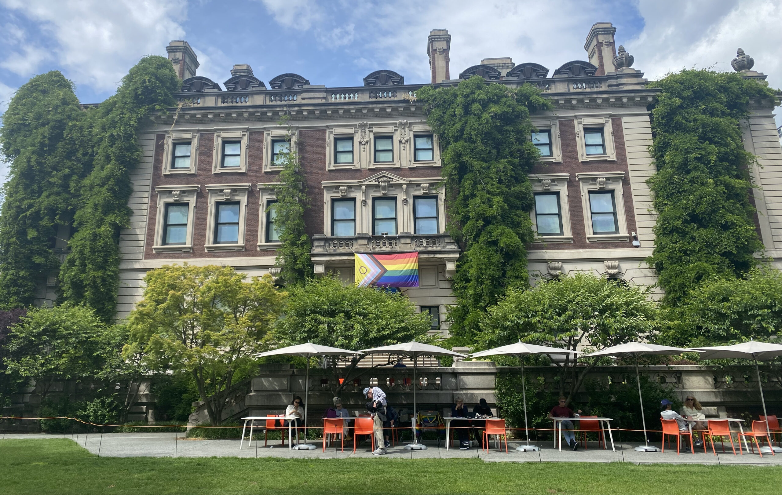 The Pride flag, of many stripes and colors, hangs from the balcony of a blocky, Georgian-style mansion with foliage covering and around it.