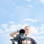 Before a pale blue sky, an individual wearing a “PRIDE” t-shirt operates a camera.