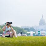 A person with green hair and rainbow–tie dye shirt sits in grass taking a photograph, with the US Capitol building in the distance.