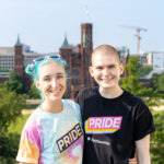 Two light-skinned individuals wearing colorful “PRIDE” t-shirts stand smiling before the brownstone Smithsonian Castle.