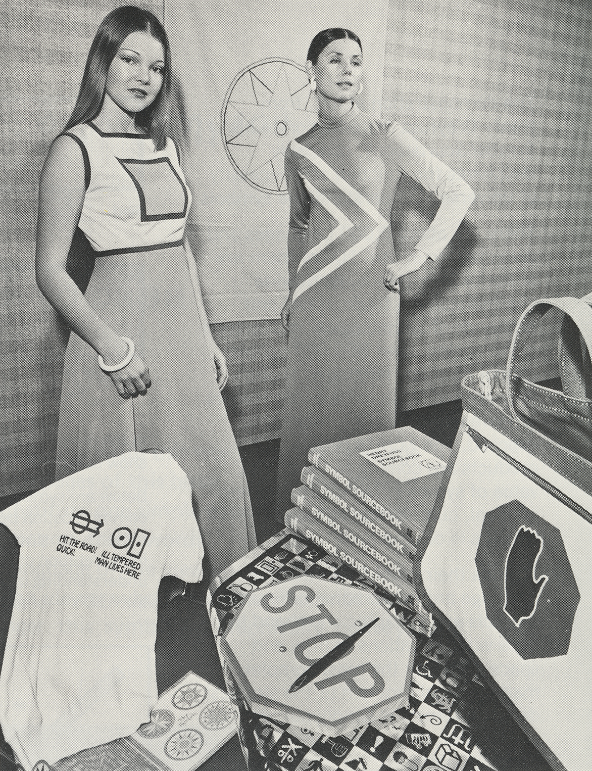 Black-and-white photograph of two women modeling long dresses with graphic detailing. In front of them is a table of merchandise related to graphic symbols.