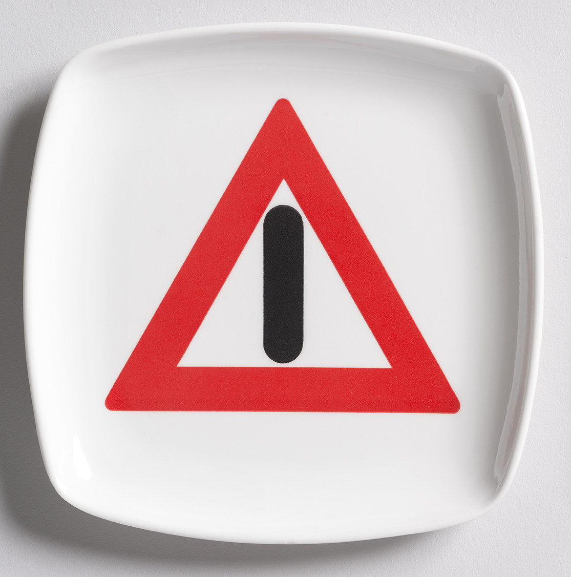 A white, rounded-square ashtray featuring a red outline of a triangle with a black vertical line in it.