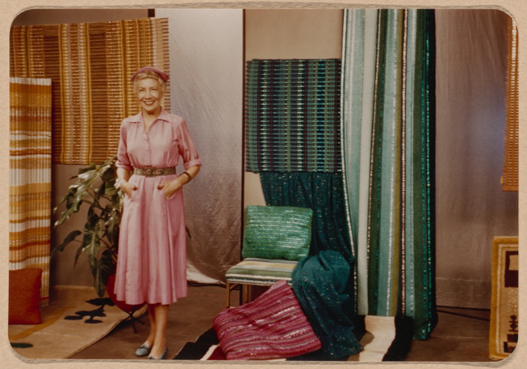 Light-skinned woman wearing pink dress and hat posing with hands in pockets in a room filled with colorful textiles.