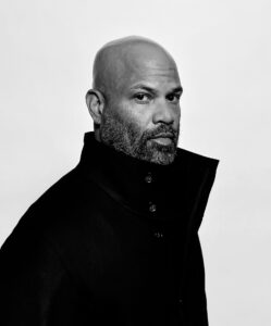 A black and white headshot image of a man with a dark jacket looking to camera.