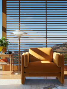 A muted yellow plush armchair sits to the right of a standing lamp in front of horizontal window shades looking outside to terrain and blue sky.