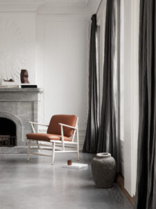 A white wooden chair with brown cushions sits next to a wall of windows with long dark grey curtains.